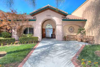 Exterior view of a single-story building with a tiled roof and an arched entrance. The building number 1350 is displayed above the entrance. There is a paved walkway leading to the entrance, bordered by green grass, shrubs, and some small trees. To the right of the entrance, there is a wooden bench and a decorative wall piece. The sky is clear and blue.