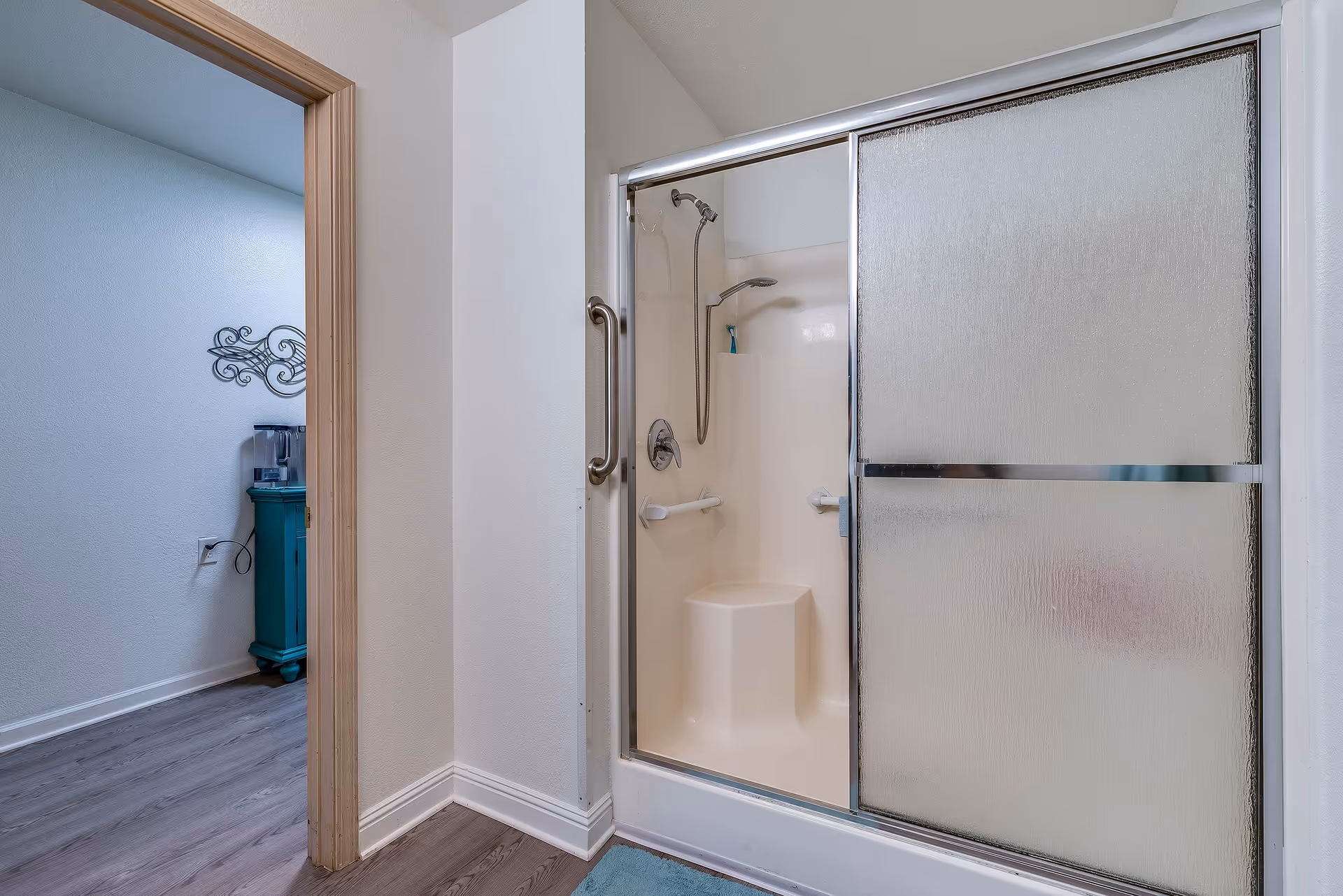 A bathroom shower with frosted sliding doors, built-in seat, grab bars and a handheld shower head, with a glimpse into an adjacent room with a teal cabinet.