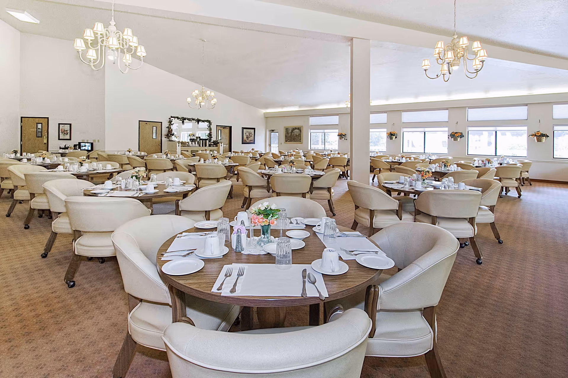 A spacious dining room with multiple round tables set with white plates, cups, silverware, and glassware. Each table has beige cushioned chairs around it and a small floral centerpiece. The room has large windows letting in natural light, beige carpeting, and white walls with framed artwork. Several chandeliers hang from the ceiling providing additional lighting.
