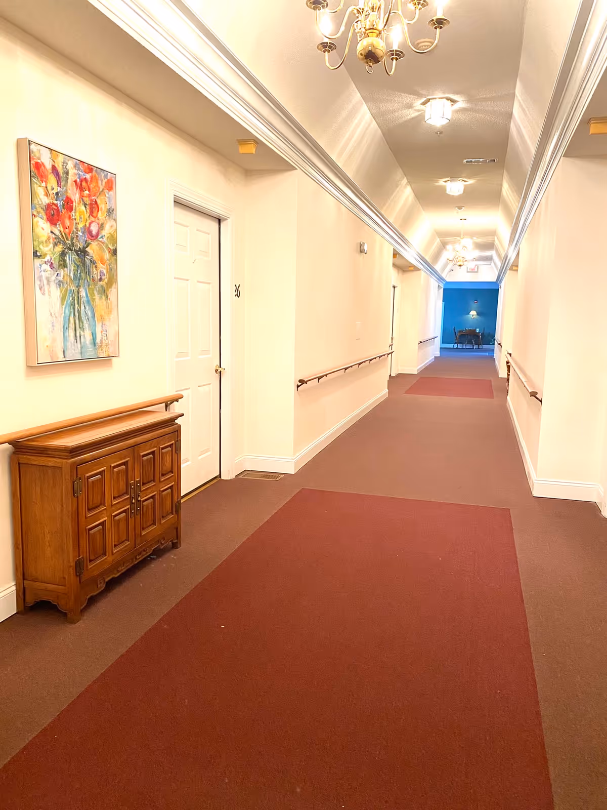 Long well-lit hallway in a senior living facility with maroon carpet runners, handrails, doors, a wooden cabinet and a floral painting.