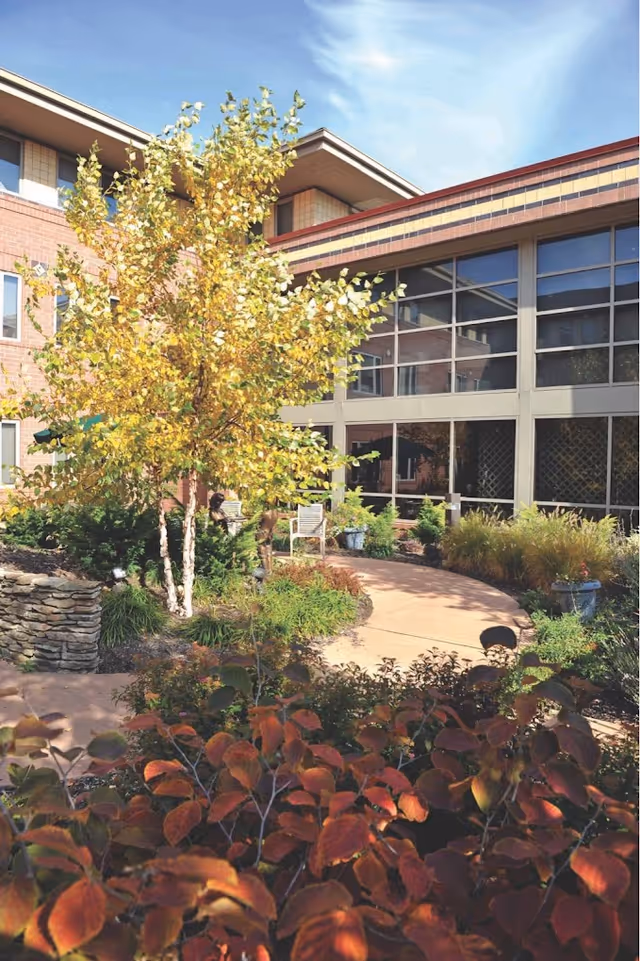 Outdoor garden area at the Daughters of Miriam Center for Nursing and Rehabilitation featuring a paved walkway, various plants and shrubs with autumn foliage, a small stone wall, and a building with large windows in the background under a clear blue sky.