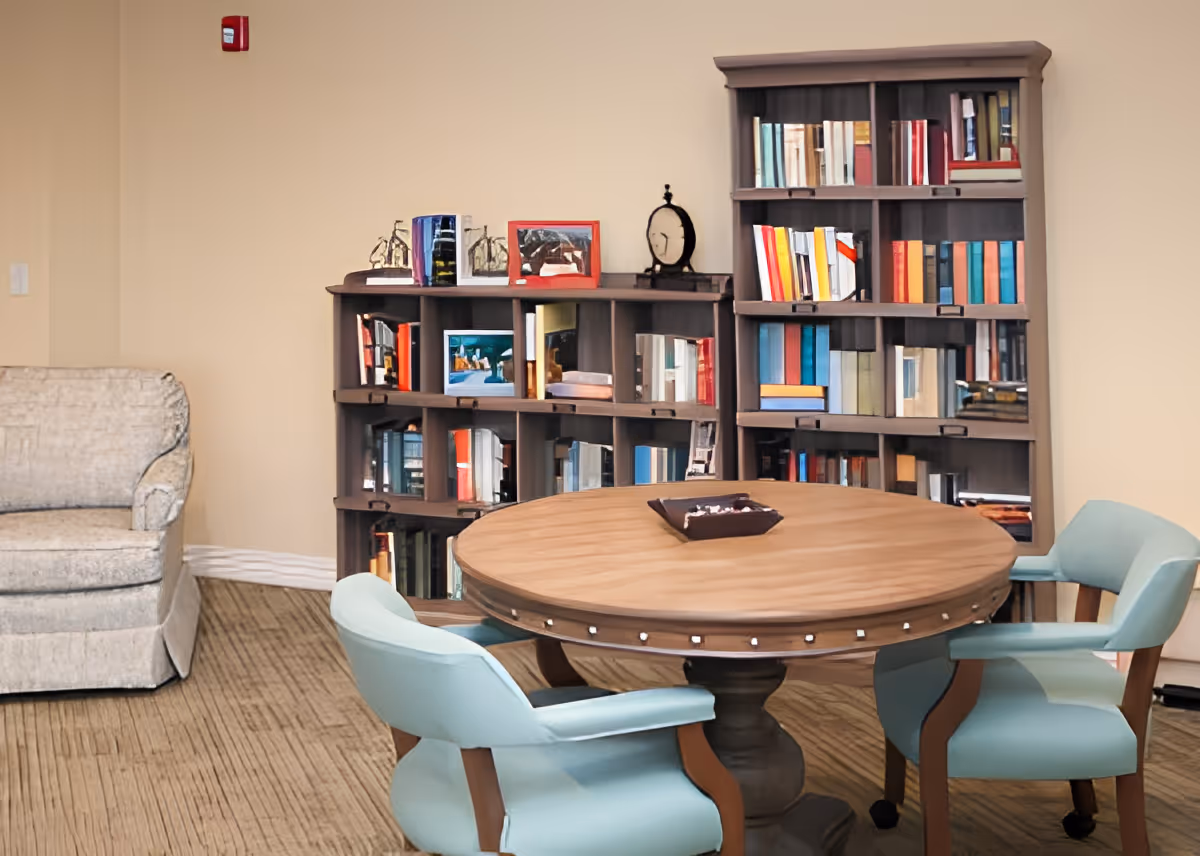 A cozy interior room with a round wooden table surrounded by four light blue cushioned chairs. Behind the table are two wooden bookshelves filled with books and decorative items. To the left, there is a beige upholstered armchair against a light-colored wall.