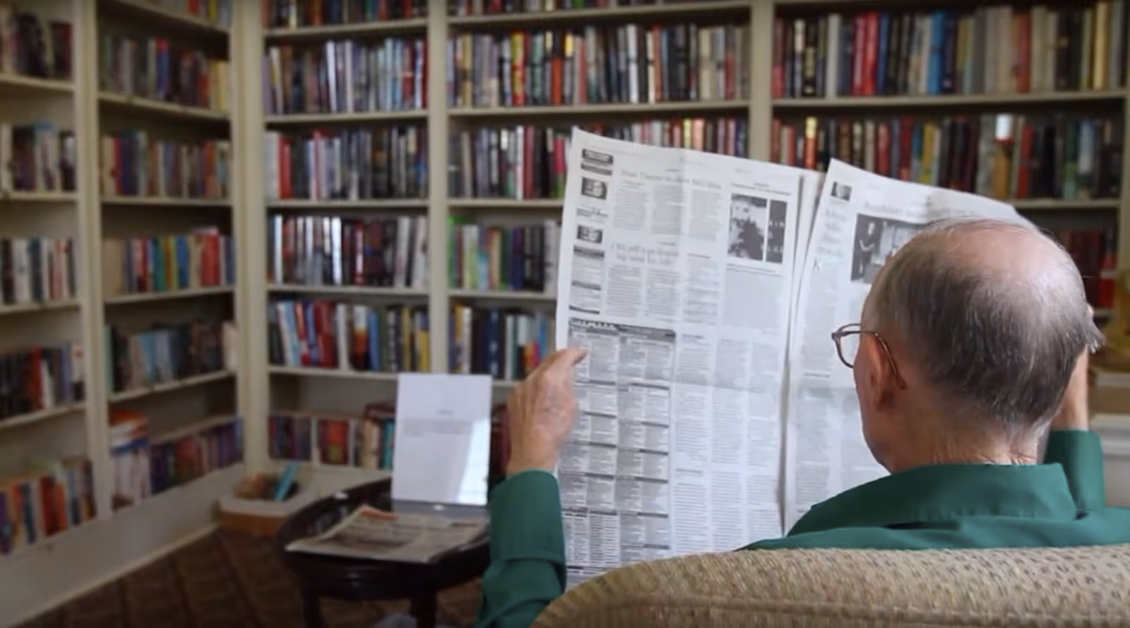 An elderly man with glasses and a green shirt is sitting in a cozy library room, reading a newspaper. Behind him are shelves filled with numerous books. A small table with more newspapers and a book stand is visible nearby.