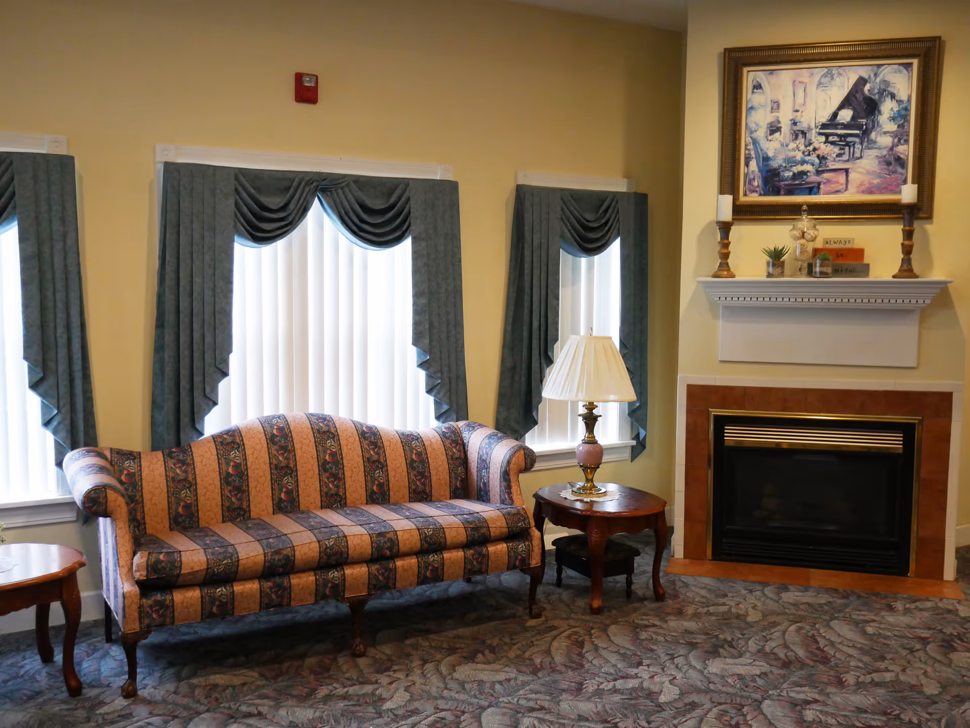 A cozy living room area with a patterned sofa, a wooden side table with a lamp, and a fireplace with a white mantel. Above the mantel is a framed painting of a piano and floral scene. The room has three windows with vertical blinds and blue valance curtains, and a patterned carpet.