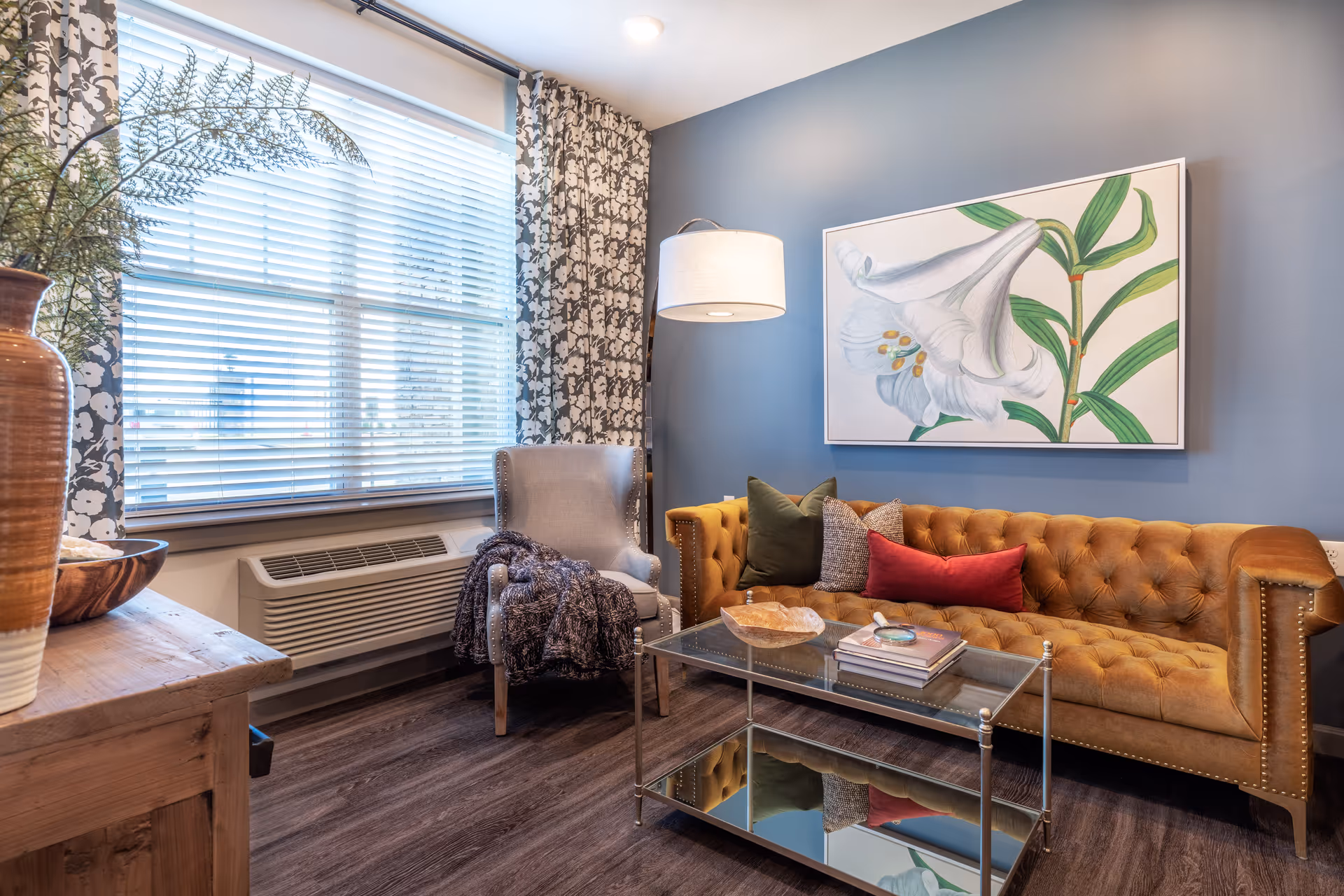 A cozy living room with a mustard yellow tufted sofa adorned with green, gray, and red pillows, a gray armchair with a dark throw blanket, a glass coffee table with books and decorative items, a large window with floral curtains, a wooden side table with a large vase and plant, and a wall-mounted painting of a white flower on a blue wall.