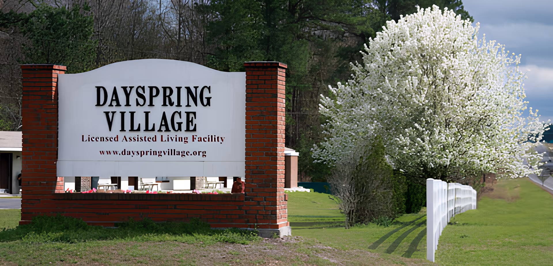 A large sign for Dayspring Village, a licensed assisted living facility, is displayed outdoors with brick pillars on either side. Behind the sign, there are trees with white blossoms and a white fence running along a grassy area.