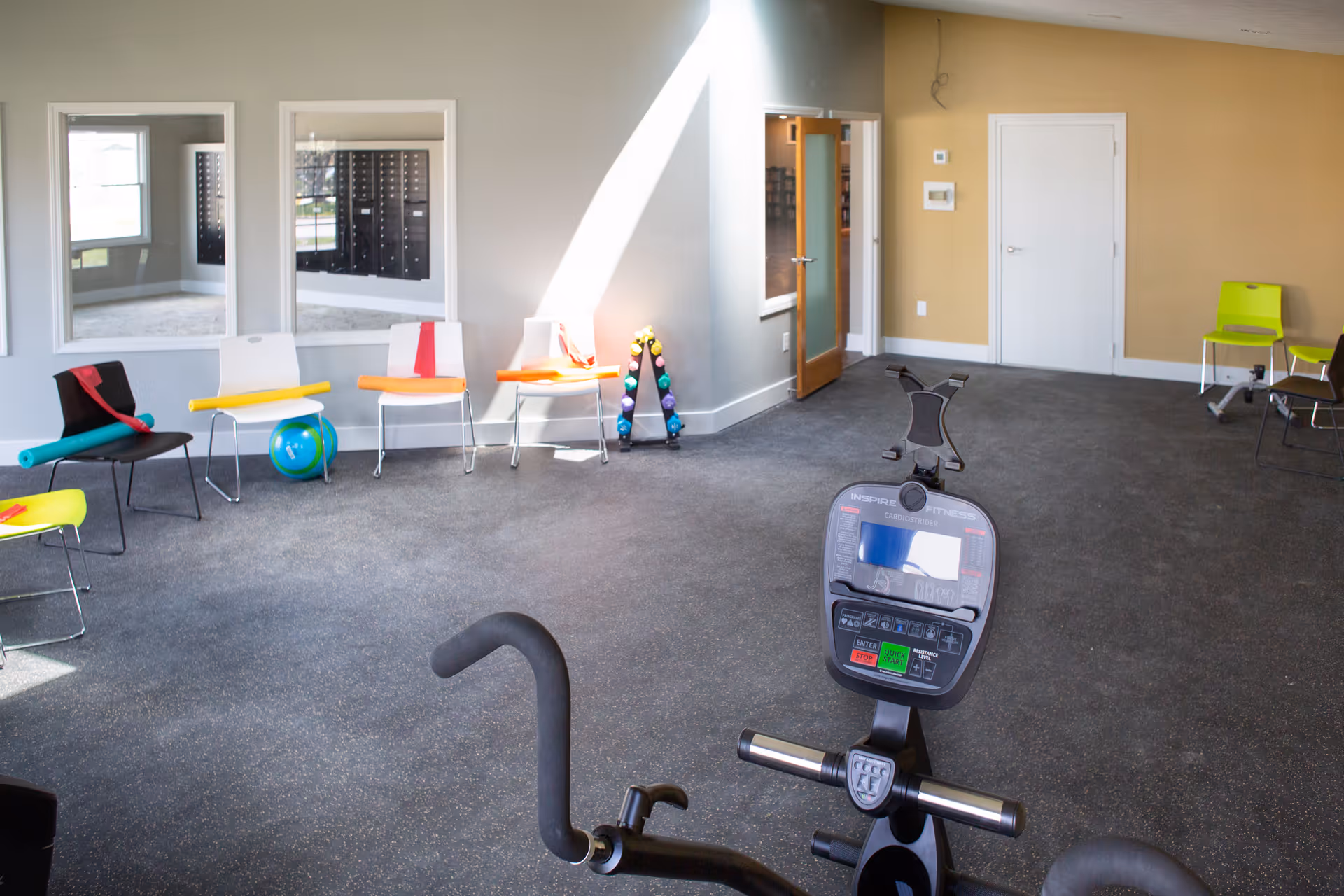 A fitness room with exercise equipment including a stationary bike in the foreground. The room has several chairs along the walls, some with exercise bands and a small exercise ball. There are large windows and mirrors on one wall, and a door leading to another room. The floor is carpeted and the walls are painted in neutral tones.