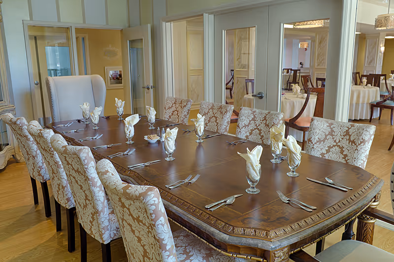 A formal dining room with a long wooden table set with glassware and folded napkins. The table is surrounded by upholstered chairs with a floral pattern. In the background, there are glass doors and additional dining tables visible in an adjoining room.