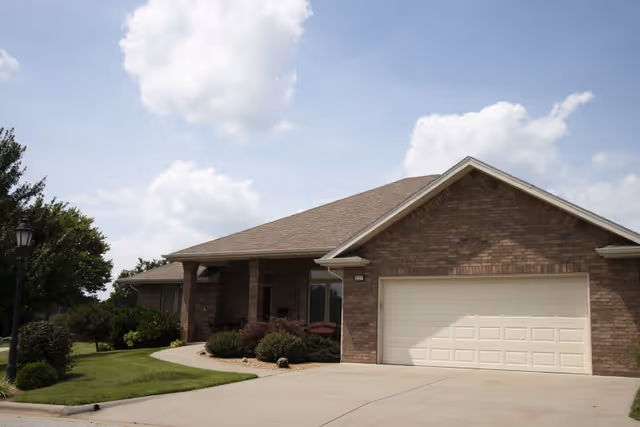 Exterior view of a single-story brick house with a two-car garage, a covered front porch, and a well-maintained lawn with shrubs and trees under a partly cloudy sky.