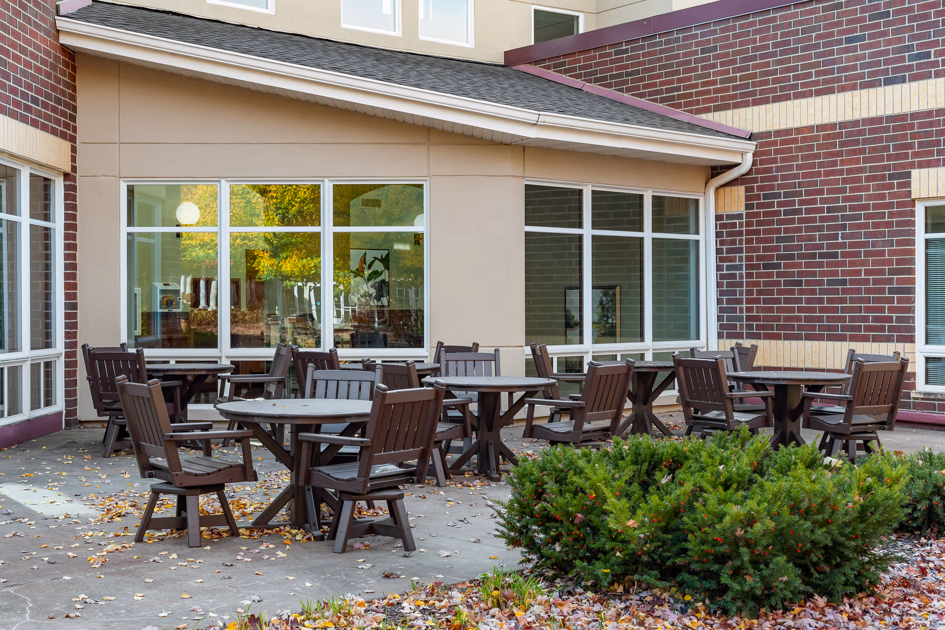 Outdoor patio area at Boutwells Landing with several round tables and wooden chairs arranged on a concrete surface. The patio is adjacent to a building with large windows and brick and beige walls. There are some green bushes and fallen autumn leaves on the ground.