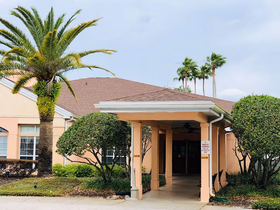 Covered entrance and porte-cochere of a peach-colored senior living building with palm trees and trimmed shrubs.
