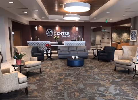 Spacious senior living lobby with armchairs, sofas, a round ottoman, and a reception desk with signage on the back wall.