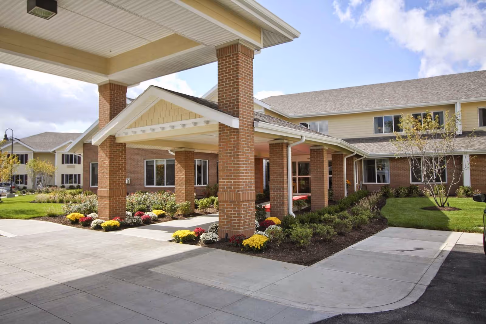 Entrance of a senior living facility with a covered drop-off area supported by brick columns, landscaped flower beds with colorful flowers, and a two-story building in the background under a partly cloudy sky.