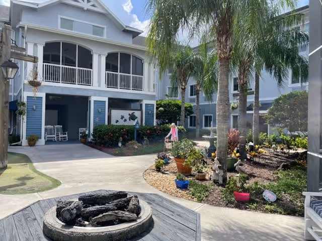 Outdoor courtyard area of a senior living facility with a fire pit in the foreground, palm trees, potted plants, and a two-story blue and white building with balconies and rocking chairs in the background under a partly cloudy sky.