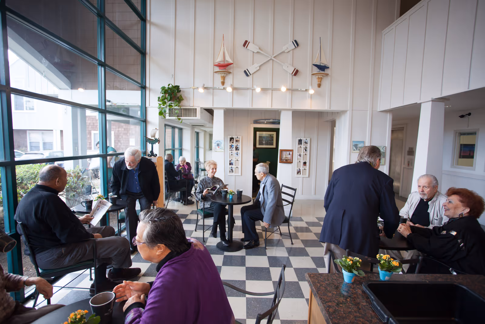A bright common area in a senior living facility with large windows on the left side. Several elderly people are sitting and standing around small tables, engaging in conversation and reading. The room has a checkered black and white tile floor, white paneled walls, and nautical decorations including two small sailboats and crossed oars mounted on the wall. Small potted plants are placed on the tables.
