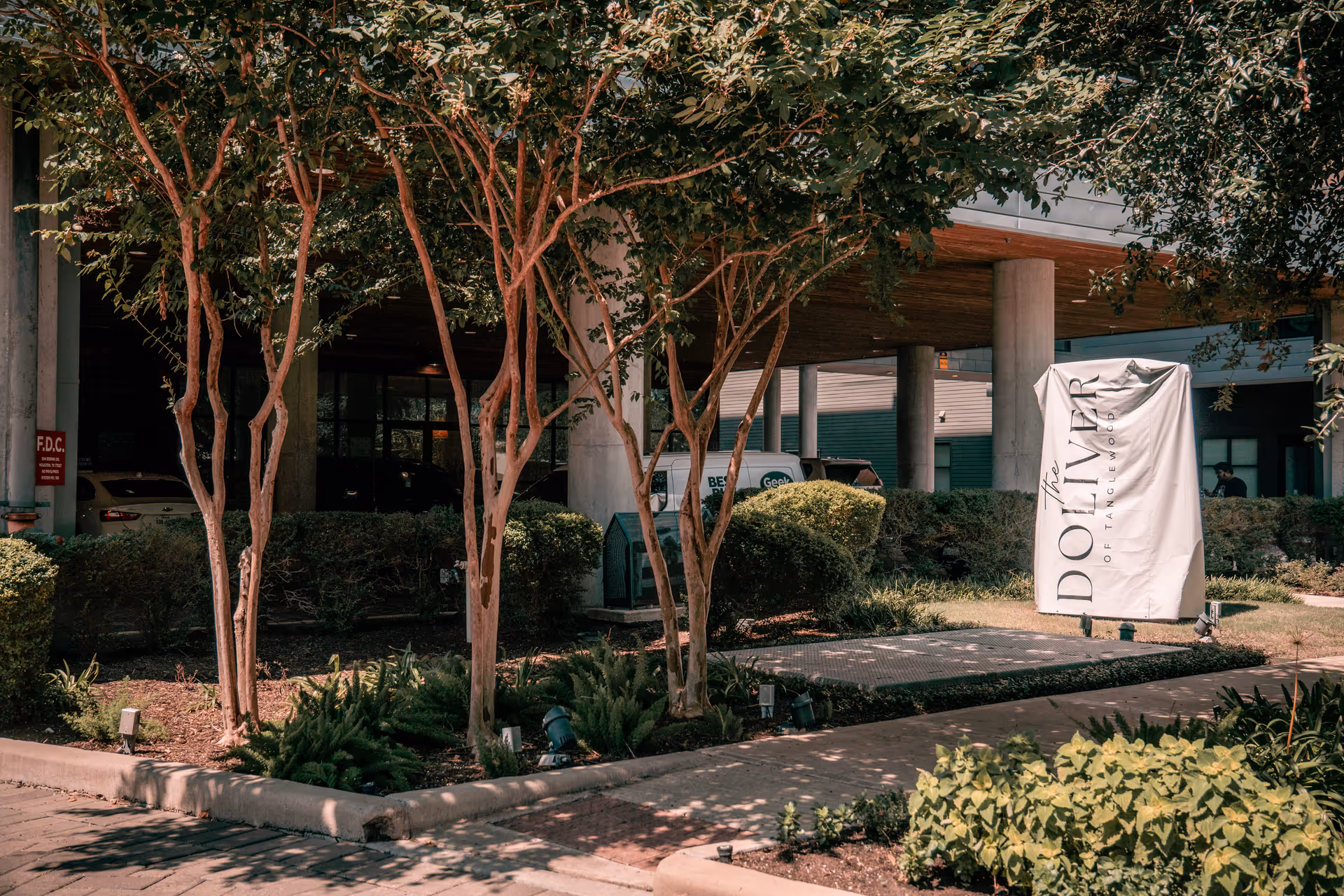 Outdoor view of The Doliver of Tanglewood facility entrance with trees, bushes, and a covered driveway area. A white sign with the facility name is prominently displayed near the sidewalk.