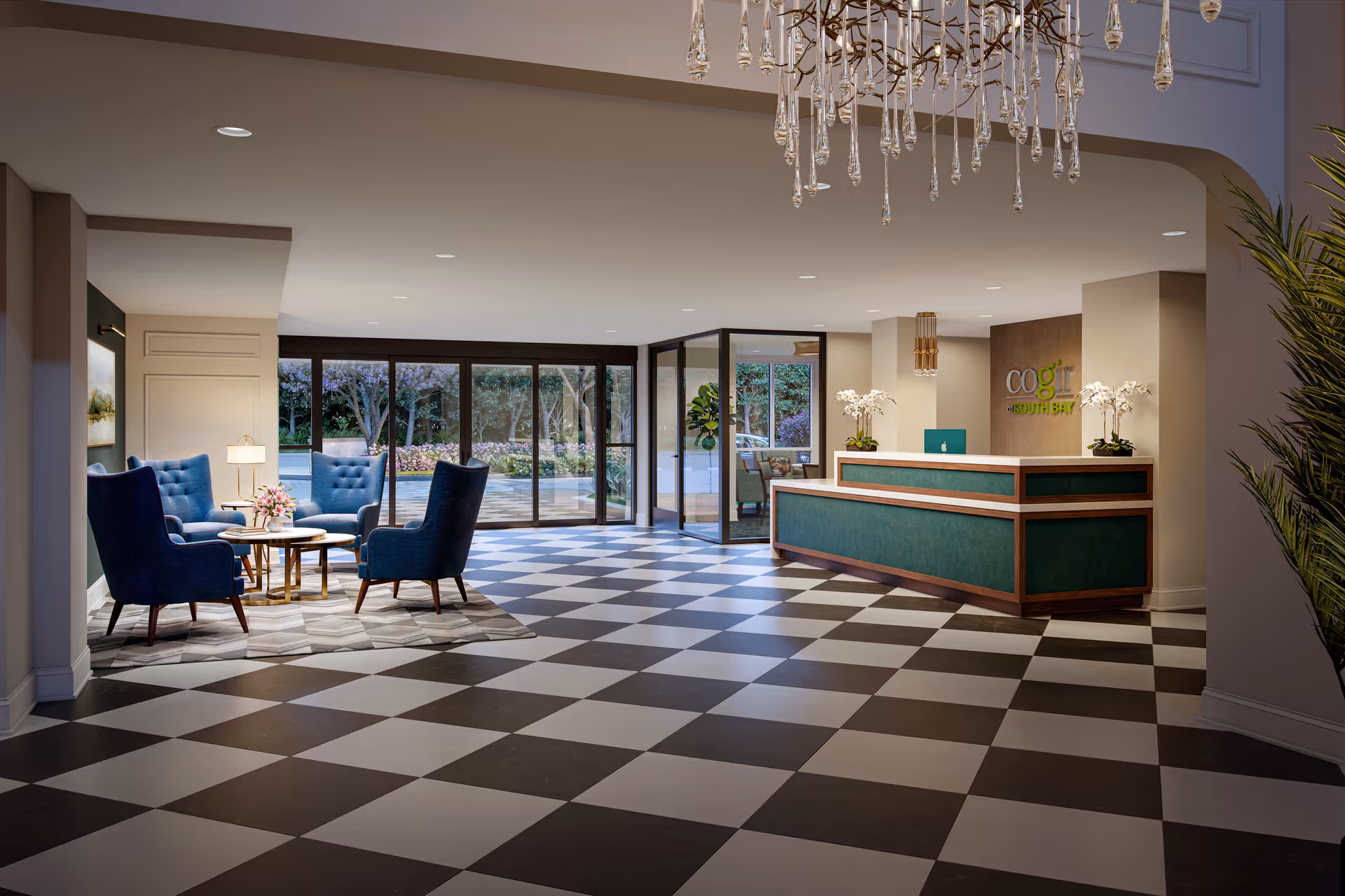 Spacious reception area of Cogir of South Bay featuring a checkered black and white floor, a green and wood reception desk with a laptop and white orchids, a seating area with four blue armchairs around a small round table with a flower arrangement, large glass doors and windows showing an outdoor garden, and a modern chandelier hanging from the ceiling.