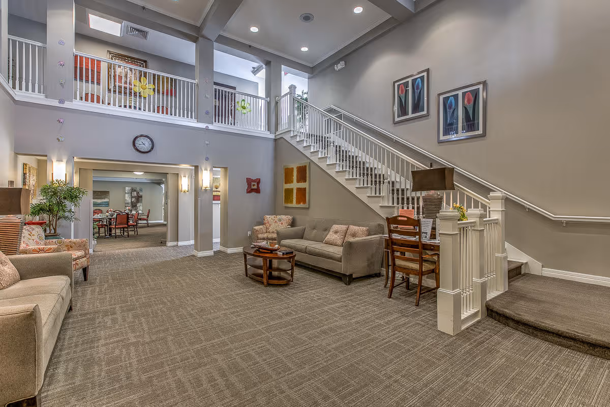 Spacious two-story assisted living lobby with sofas, chairs, a central coffee table, and a staircase leading to an upper balcony with a dining area visible through an archway.