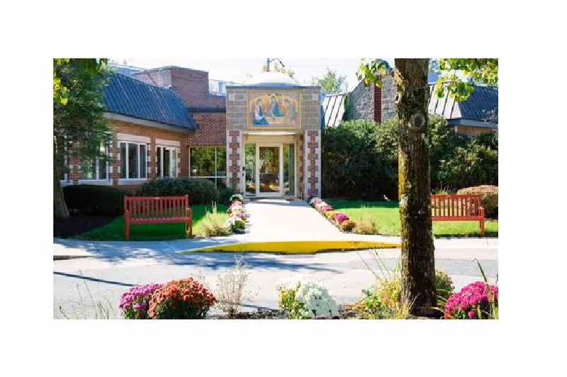 Front entrance of a nursing facility with a central glass doorway, a walkway lined with flowers, red benches, and landscaping.