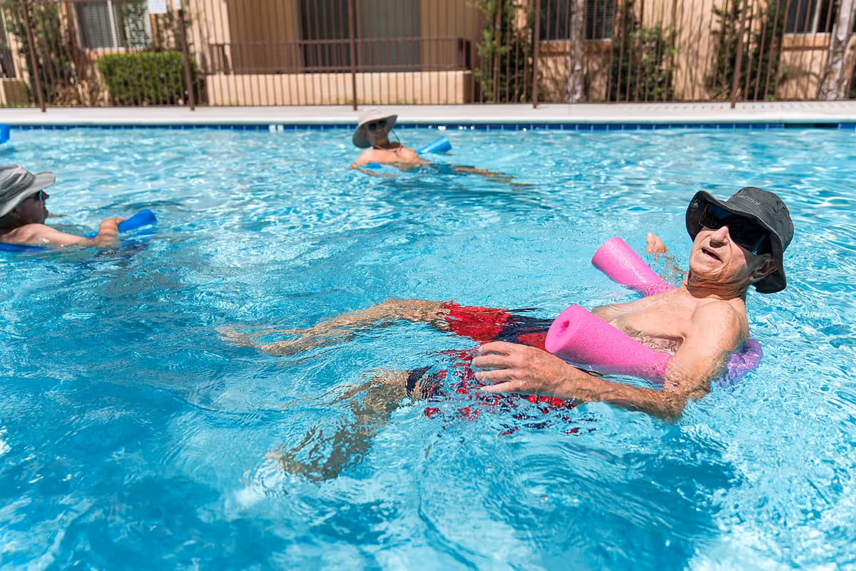 Three elderly men wearing hats and sunglasses relaxing and floating in a swimming pool using pool noodles on a sunny day.