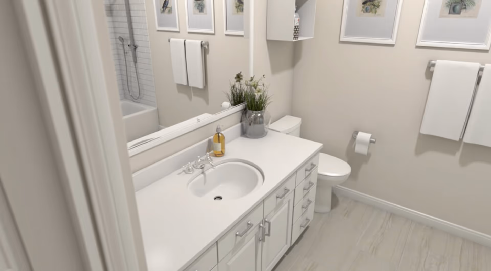A clean and modern bathroom with a white vanity featuring a sink, silver faucet, and multiple drawers and cabinets. There is a large mirror above the vanity, a toilet next to it, and a bathtub with a showerhead visible in the background. Two white towels hang on a towel rack on the wall, and three framed pictures are displayed above the towels. A small plant in a glass vase and a soap dispenser are placed on the vanity countertop.