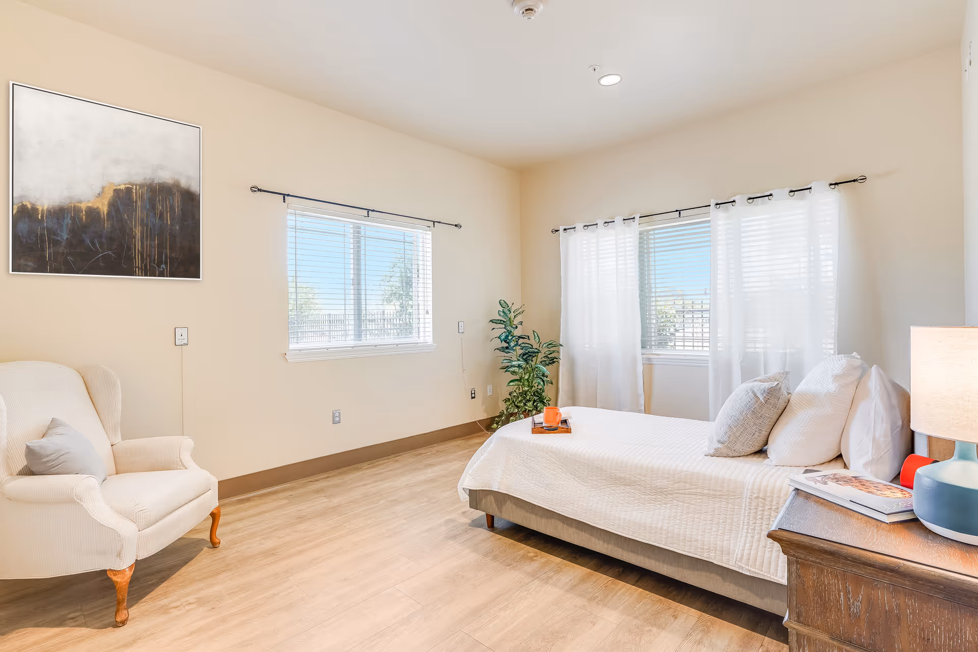 Bright, simply furnished bedroom with a single bed, armchair, nightstand with a lamp, and windows with sheer curtains.