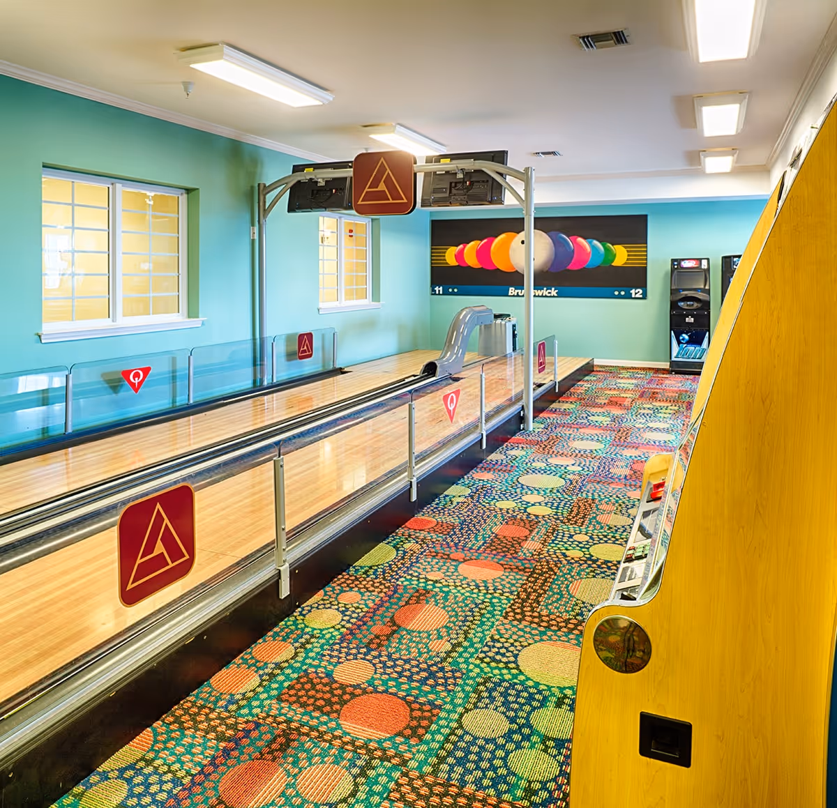 Indoor bowling alley with two lanes, colorful patterned carpet, teal walls, two windows, and a large colorful bowling ball mural on the far wall. There is an arcade game machine on the right side.