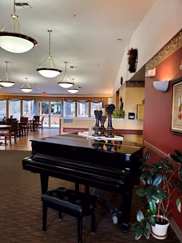 Interior view of a senior living facility common area featuring a black grand piano with a matching bench in the foreground. The piano top holds decorative items including three black ornamental pieces and a tray. In the background, there are several wooden tables and chairs arranged near large windows with blue curtains, allowing natural light to fill the room. The ceiling has multiple round pendant lights, and the walls are decorated with framed artwork and plants.