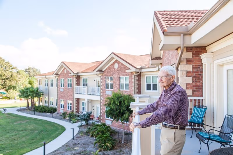 An elderly man stands on a balcony overlooking the landscaped courtyard and brick exterior of a senior living building.
