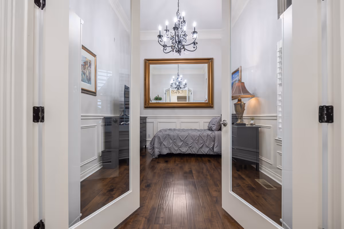 View through open glass French doors into a bedroom with a bed, chandelier, large framed mirror, nightstand with lamp, and hardwood floors.