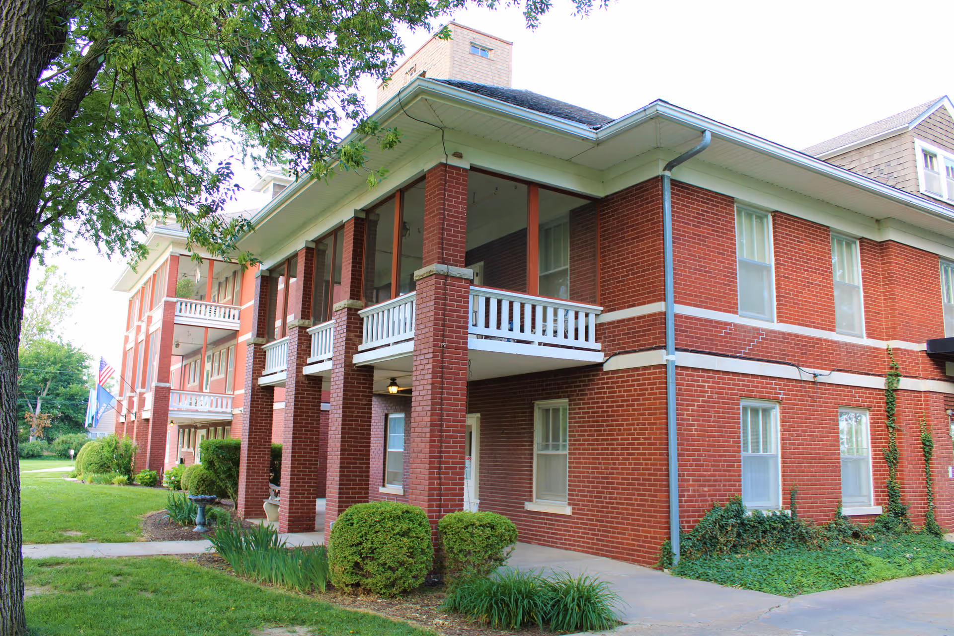Red brick two-story building with white balconies, columns, and surrounding lawn and trees.