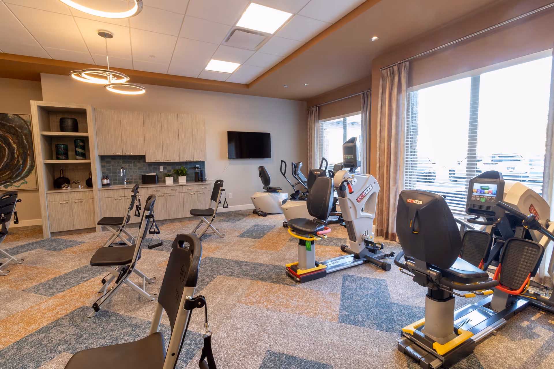 A bright exercise room with several stationary recumbent bikes and folding chairs spaced out on a patterned carpet. The room has large windows with curtains letting in natural light, a wall-mounted TV, and a kitchenette area with cabinets and a sink. Modern circular ceiling lights hang from the ceiling.