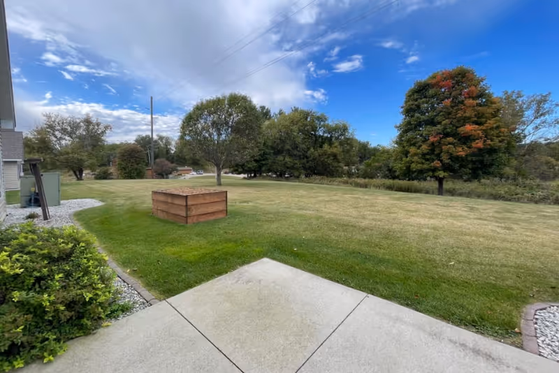 Open grassy yard with a concrete patio, a wooden planter box, and trees under a partly cloudy sky.