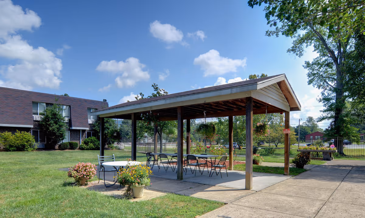 Outdoor covered pavilion with tables and chairs set on a concrete patio surrounded by green grass and potted flowers. In the background, there is a two-story building with windows and trees under a blue sky with scattered clouds.