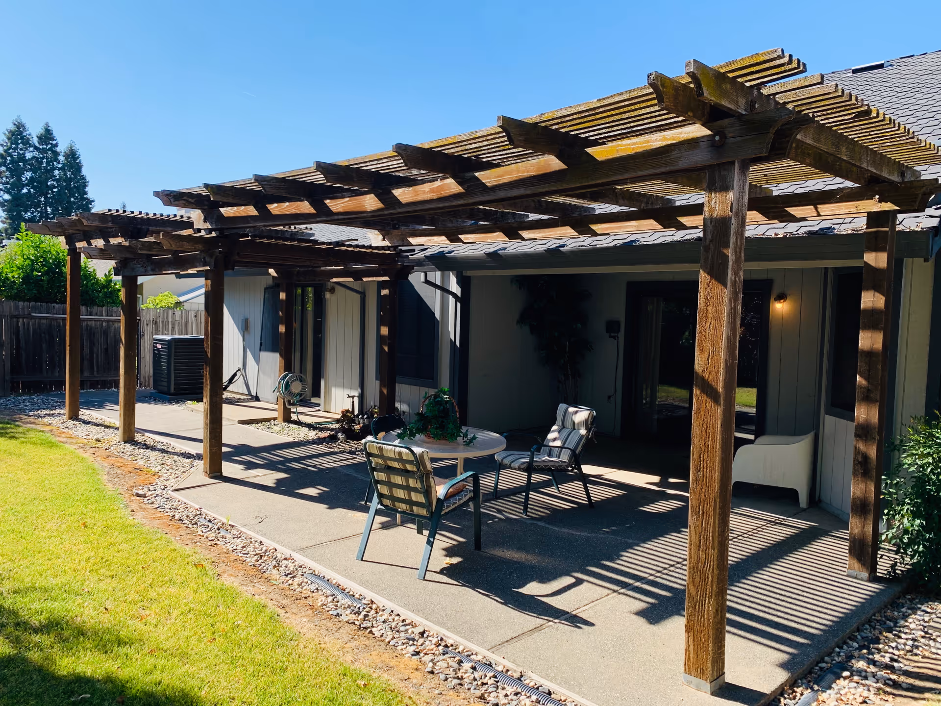 Outdoor patio area with a wooden pergola attached to a building. The patio has a small round table with two chairs and a white armchair. The ground is paved with concrete and bordered by small rocks. There is a grassy lawn adjacent to the patio and a wooden fence in the background.