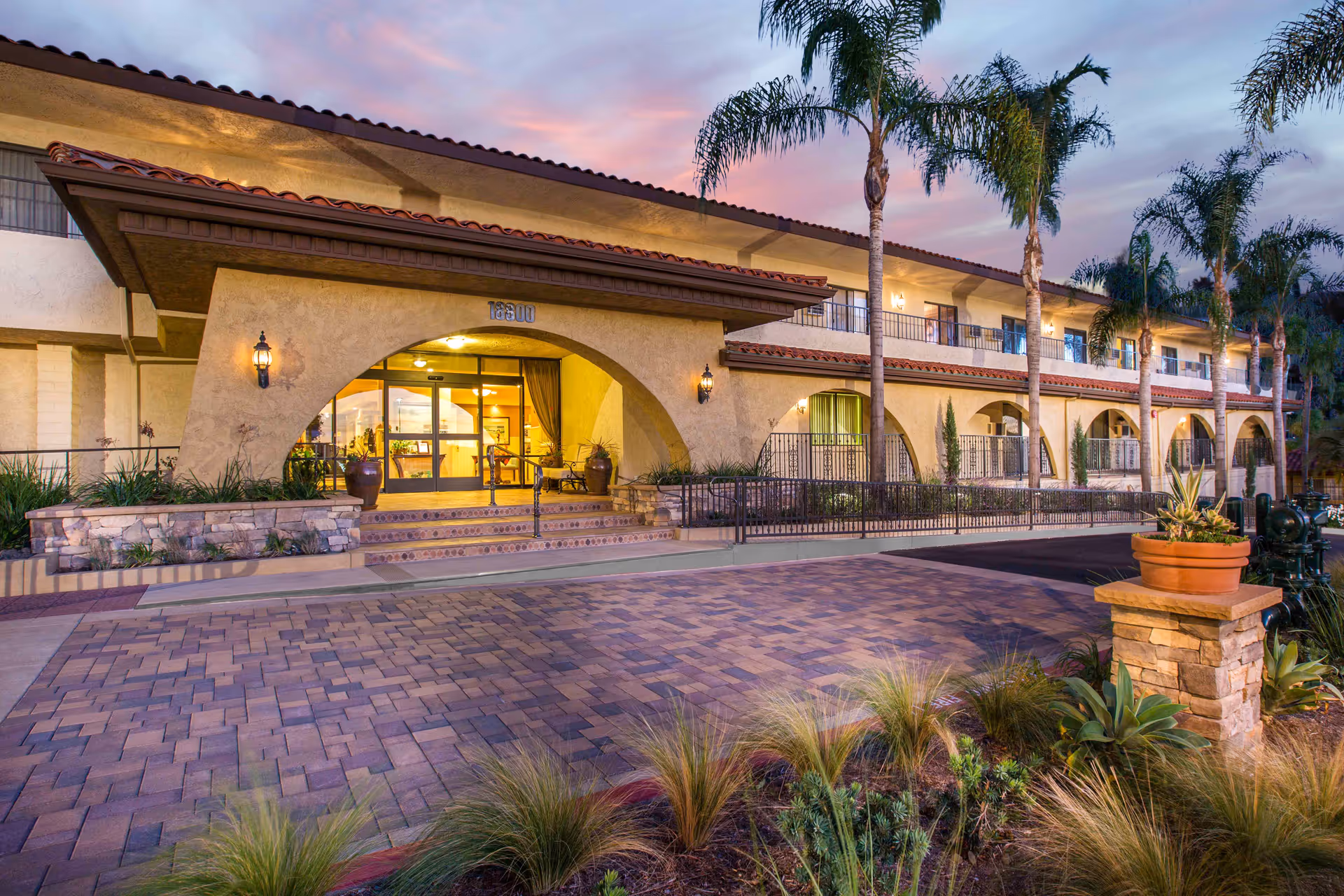 Front entrance of a two-story Mediterranean-style senior living building with arched walkways, palm trees, and a paved driveway at dusk.
