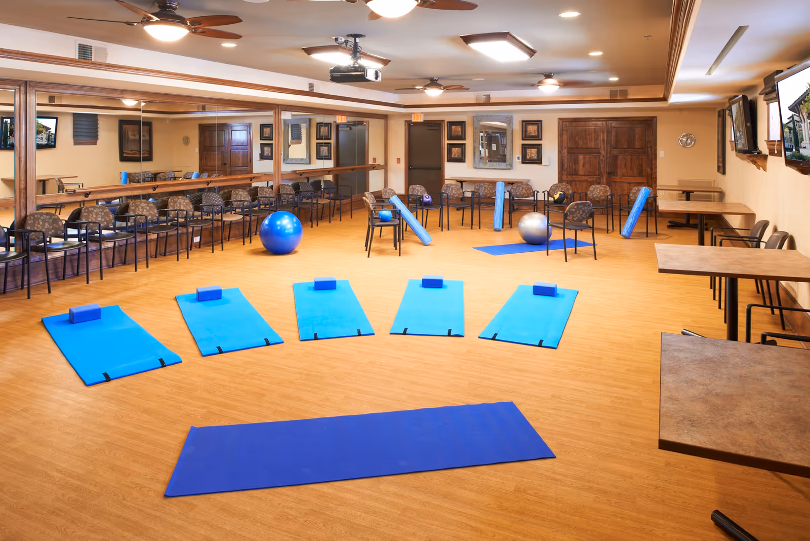 A spacious exercise room with wooden flooring and beige walls, featuring five blue exercise mats with blocks arranged on the floor, several exercise balls, foam rollers, and chairs lined up against the mirrored wall. There are tables and chairs along the right side of the room and ceiling fans above.