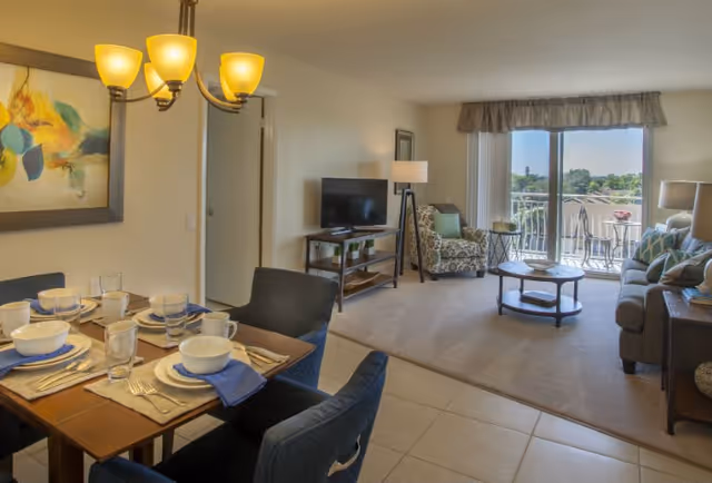 A bright and spacious living and dining area in a senior living facility. The dining table is set with plates, bowls, glasses, and silverware, with blue napkins. The living room features a sofa, an armchair with a patterned fabric, a coffee table, a TV on a stand, and a floor lamp. Large sliding glass doors lead to a balcony with outdoor seating and a view of greenery outside.