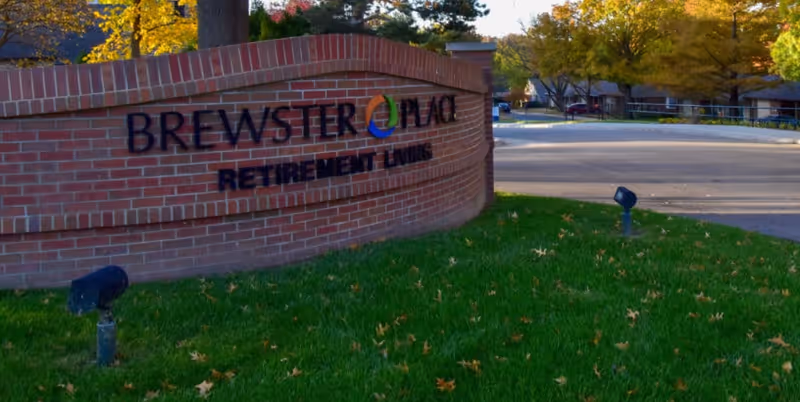 Brick sign for Brewster Place Retirement Living on a grassy area with trees and a driveway in the background during autumn.