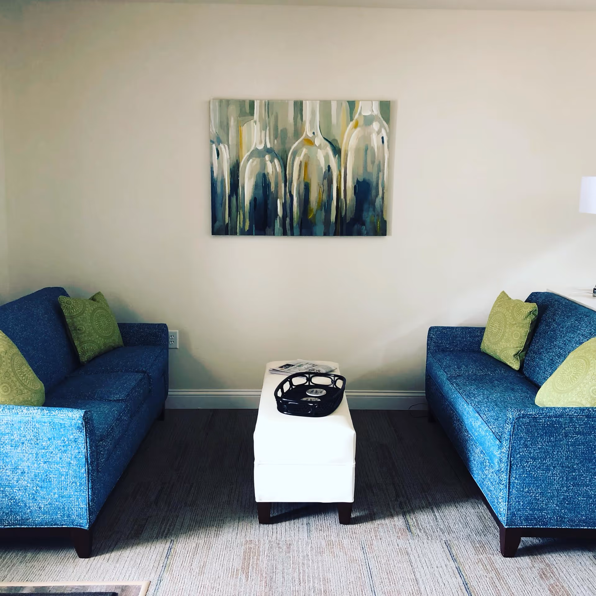 A small sitting area with two blue upholstered sofas facing each other, each with two green patterned throw pillows. Between the sofas is a white ottoman with a black tray on top. A painting of abstract glass bottles hangs on the beige wall behind the ottoman.