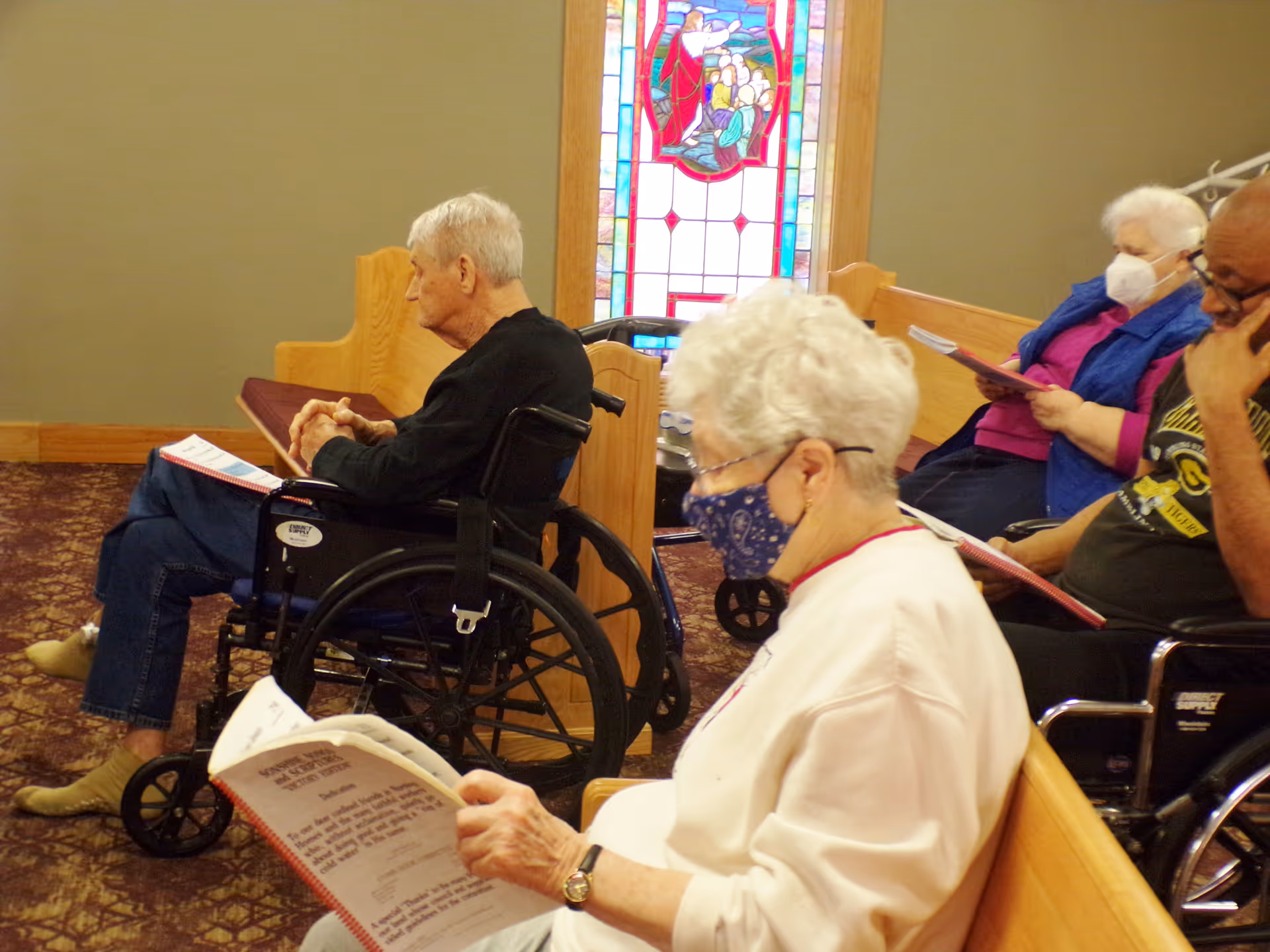 Several elderly individuals, some in wheelchairs, seated on wooden pews inside a room with a stained glass window. They are wearing face masks and reading from booklets.