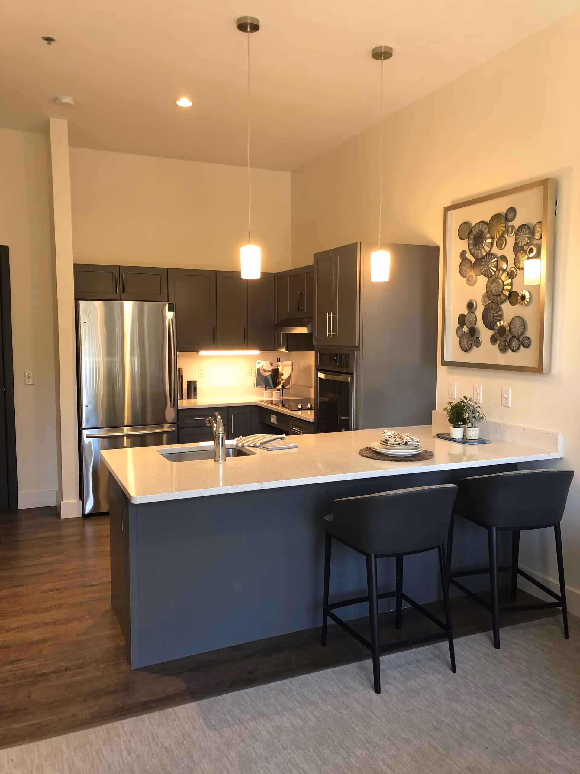 Modern kitchen with gray cabinets, stainless steel refrigerator, built-in oven, and a white countertop island with a sink and two black bar stools. Two pendant lights hang from the ceiling above the island. A decorative wall art piece is mounted on the wall to the right.