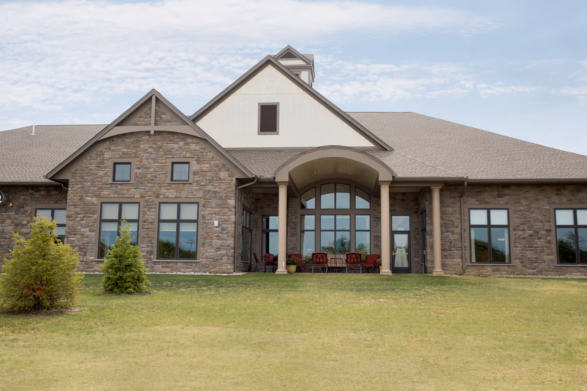 Exterior view of a single-story stone building with large windows and a covered patio area furnished with red cushioned chairs and tables, set against a partly cloudy sky and a grassy lawn with small trees.