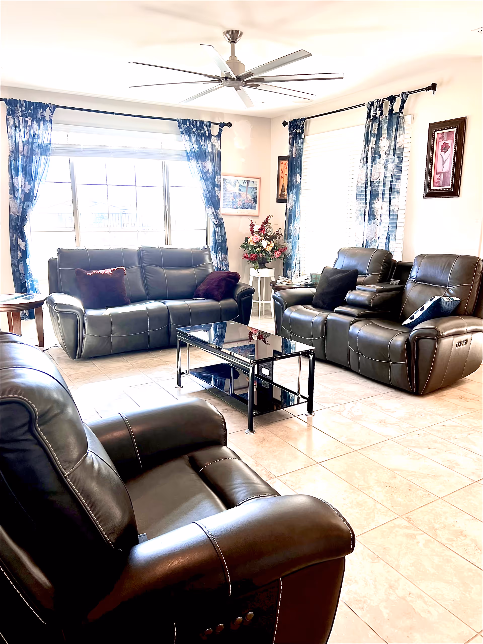 A bright living room with large windows covered by blue floral curtains. The room features dark leather recliner chairs and a matching sofa with dark throw pillows. A black glass coffee table is centered on the tiled floor. There are framed pictures on the walls and a floral arrangement on a small white table in the corner. A large ceiling fan is mounted on the white ceiling.