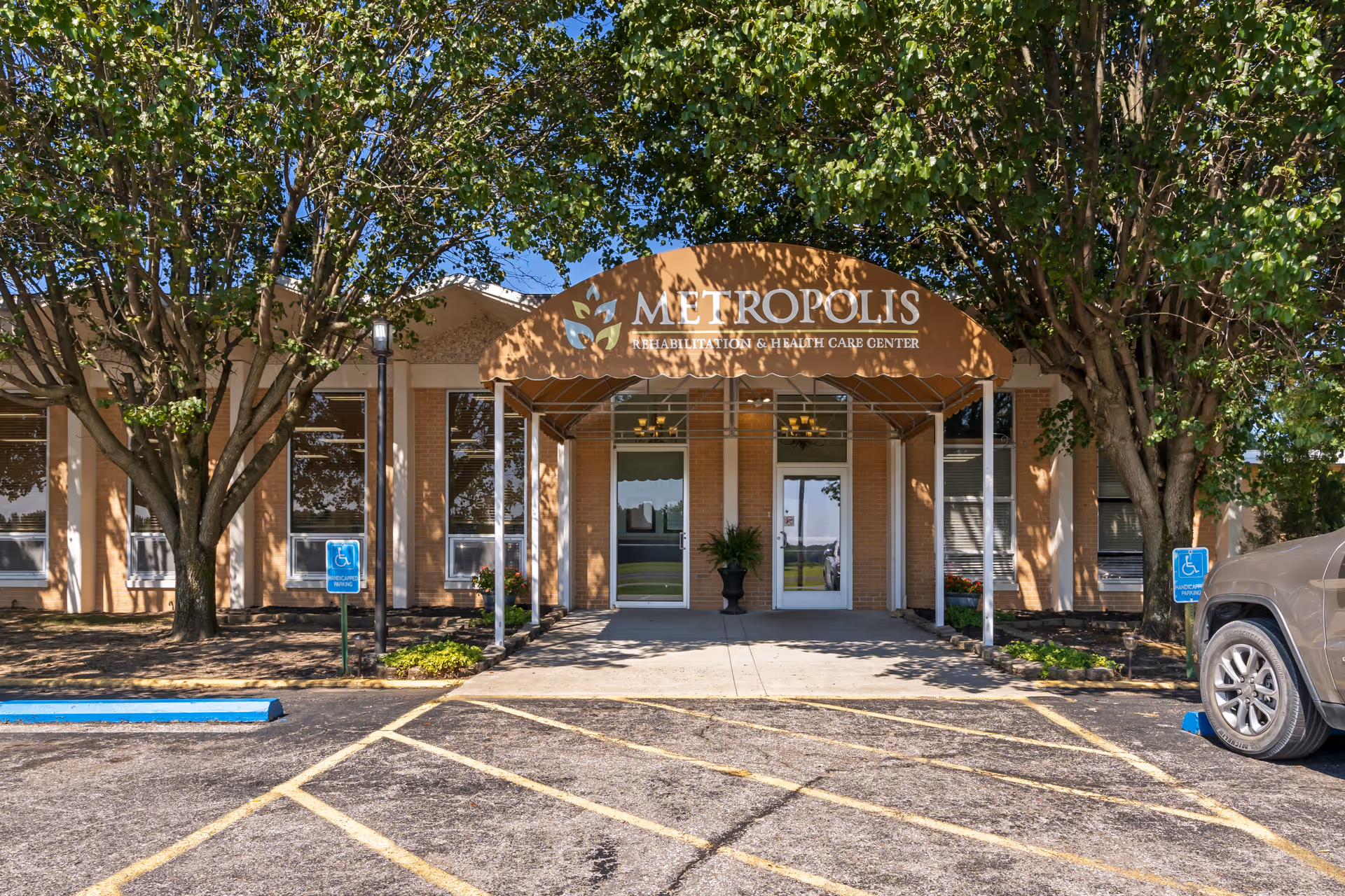 Front exterior view of Metropolis Rehabilitation & Health Care Center with a brown awning displaying the facility name, flanked by trees and handicap parking spaces in front.