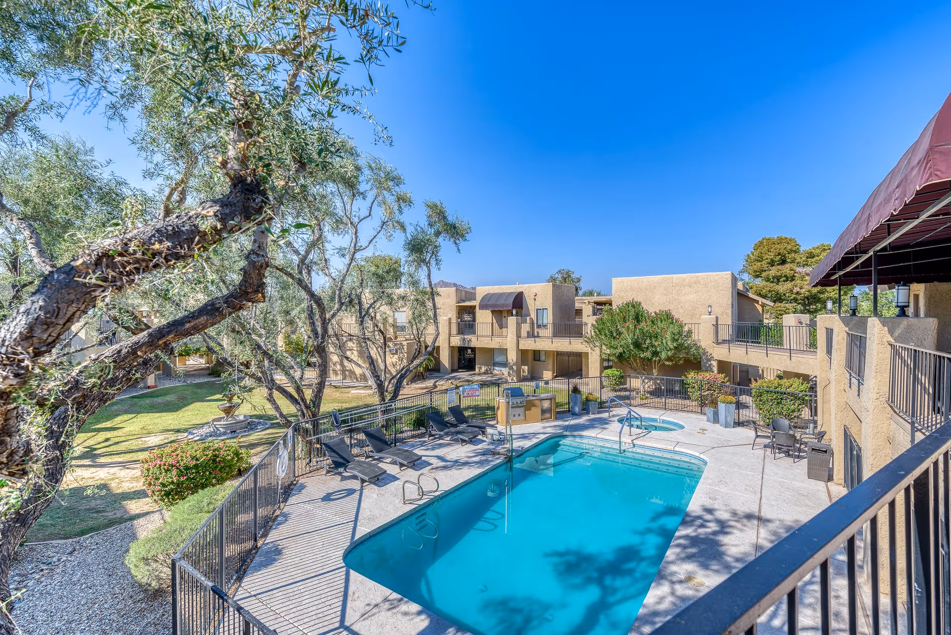 Outdoor view of a senior living facility courtyard featuring a swimming pool and a hot tub surrounded by a safety fence. There are lounge chairs and tables with umbrellas around the pool area. The courtyard is landscaped with trees, bushes, and a grassy area. Beige two-story buildings with balconies and awnings surround the courtyard under a clear blue sky.