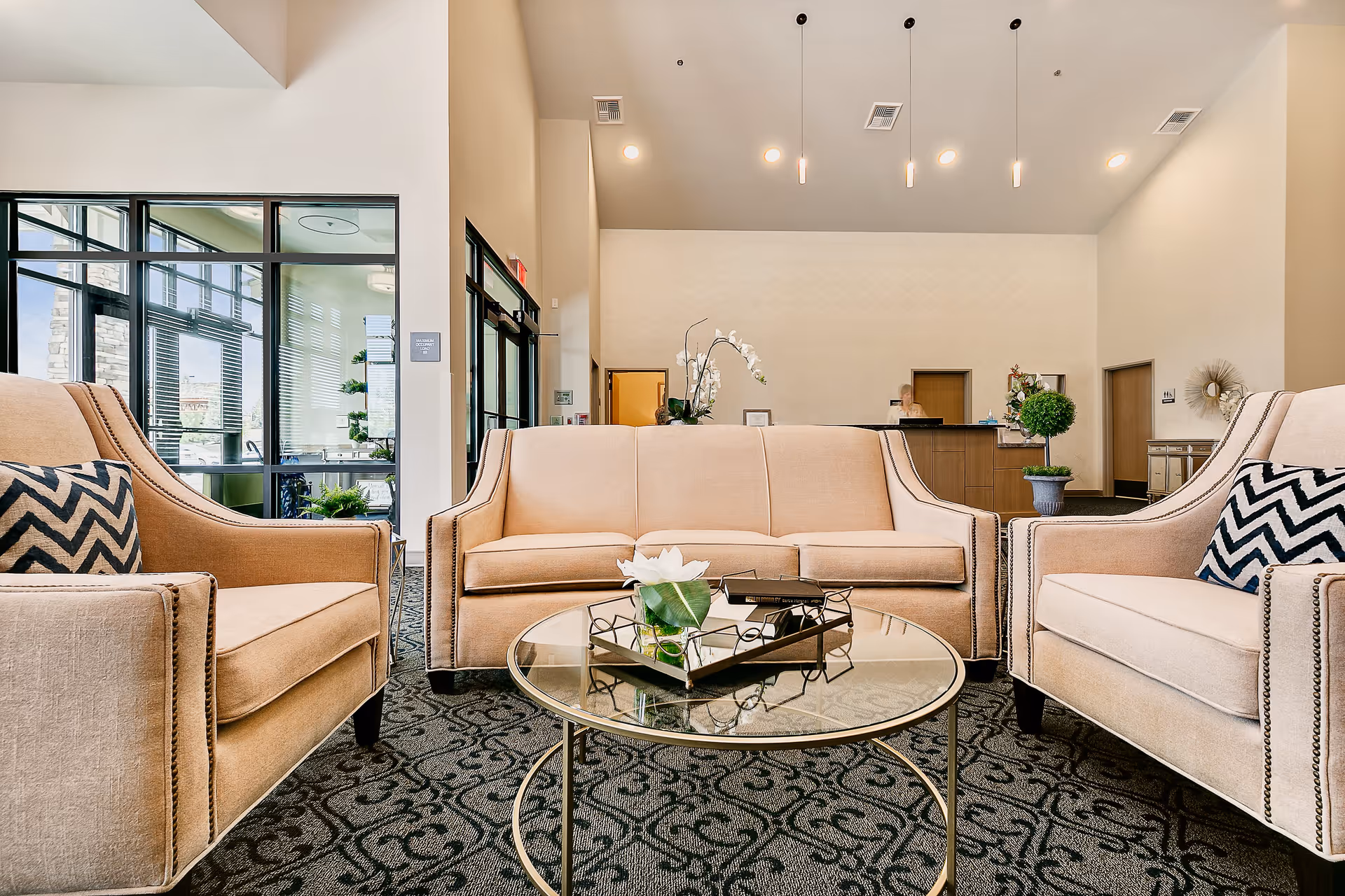 A bright and spacious senior living facility lobby with beige upholstered armchairs and a matching sofa arranged around a round glass coffee table. The table has a decorative tray with a white flower and books. The background shows a reception desk with a staff member, potted plants, and large windows letting in natural light.