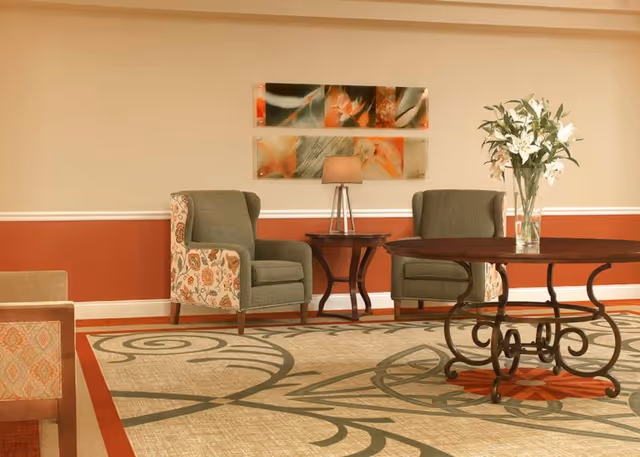 A cozy sitting area with two upholstered armchairs featuring floral patterned sides, a small wooden side table with a lamp, and a round wooden table with a decorative wrought iron base holding a vase of white lilies. The room has a patterned carpet, a two-tone wall with a chair rail, and two rectangular floral art pieces hanging on the wall.