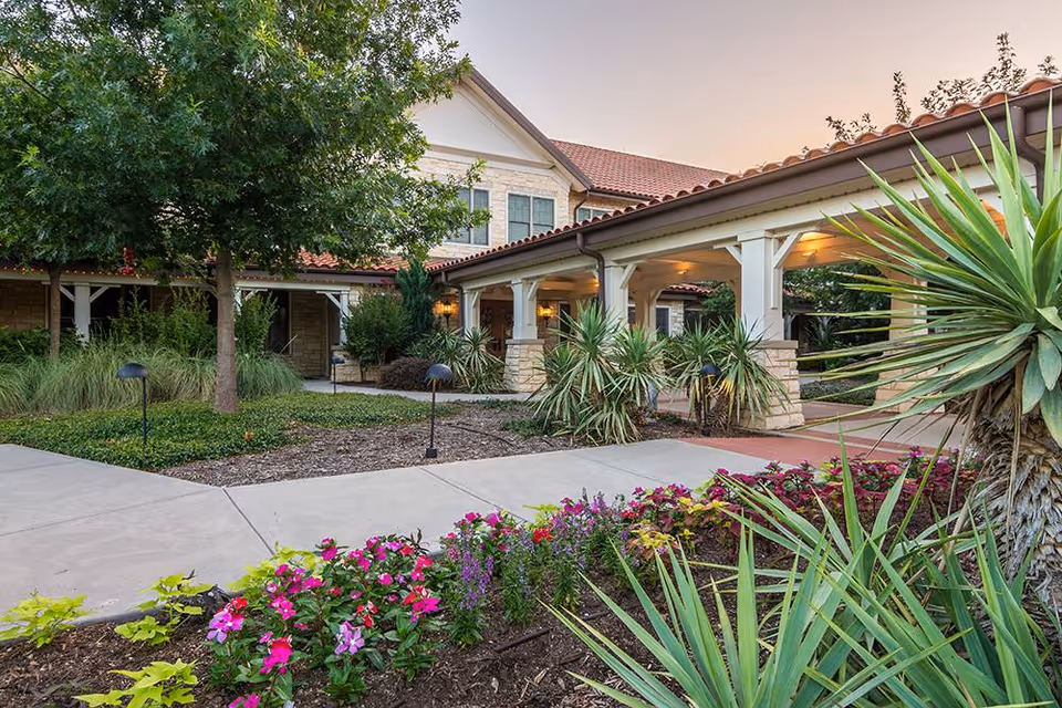 Exterior view of a senior living facility with a covered walkway supported by white columns and stone bases. The building has a tiled roof and beige stone walls. In the foreground, there is a landscaped garden with colorful flowers, green shrubs, and a tree. The sky is clear with a soft evening light.
