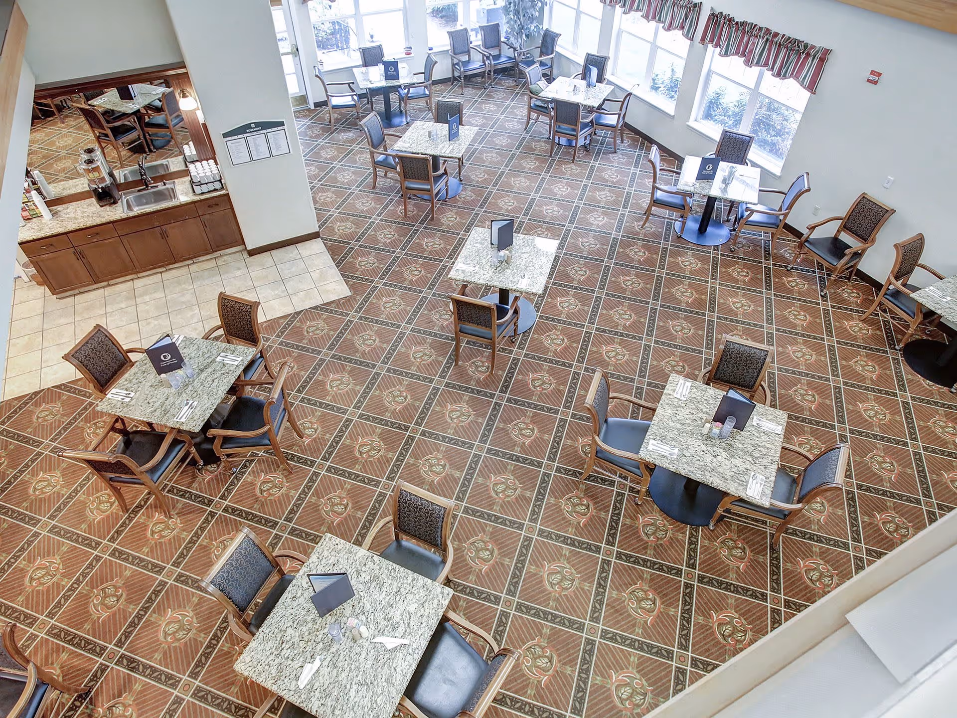 A dining room in a senior living facility with multiple square tables, each surrounded by four chairs. The tables have granite tops and are set with menus, napkins, and utensils. The floor has a patterned tile design, and large windows with valances allow natural light to fill the room. There is a small counter area with a sink and beverage dispenser in one corner.